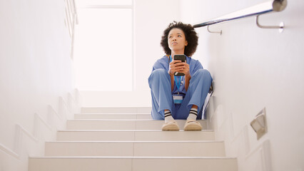 Stressed Female Nurse Wearing Scrubs Browsing On Mobile Phone Sitting On Stairs In Hospital