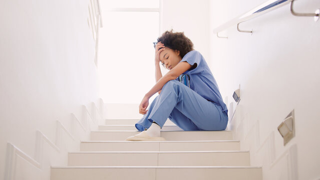 Stressed And Tired Female Nurse Wearing Scrubs Sitting On Stairs In Hospital