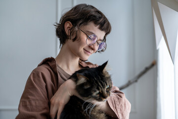 Contented woman hugs fluffy cat at home near window. Tenderness, love for pets. Happy young female...