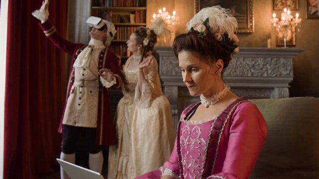 Medium shot of young Caucasian lady actress in pink Victorian corset dress, elaborate hairdo with feathers, flowers browsing online on laptop, relaxing between shooting scenes for historical drama