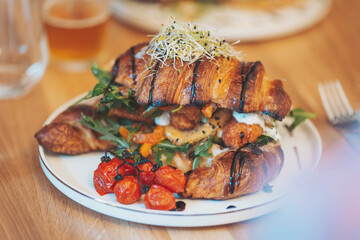 Savory croissant sandwich piled high with fresh greens, grilled vegetables, and sauces on a rustic wooden table during brunch hour