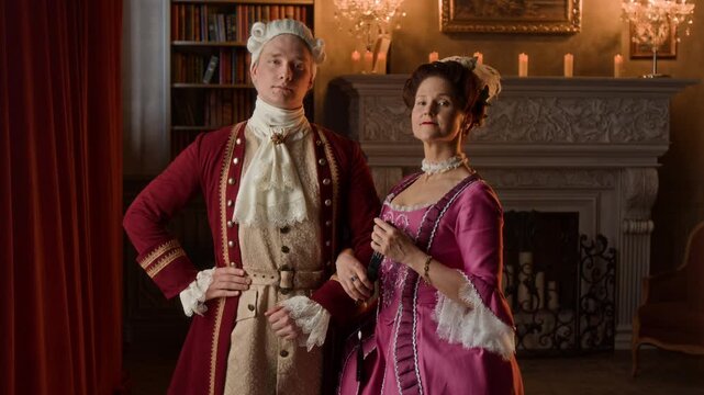 Medium portrait of young Caucasian noble woman in pink frilly dress, with elaborate hairdo, and gentleman in wig and red surcoat, standing in mansion hall, holding hand, looking at camera