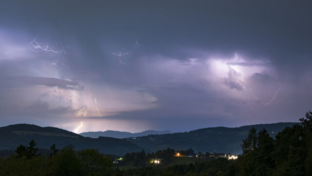 View of dramatic lightning bolts illuminating the night sky above rolling hills and distant structures, Graz, Styria, Austria.