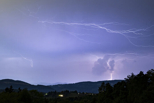 View of jagged lightning bolts electrifying the twilight sky above the dark rolling hills and scattered lights in Graz, Styria, Austria.