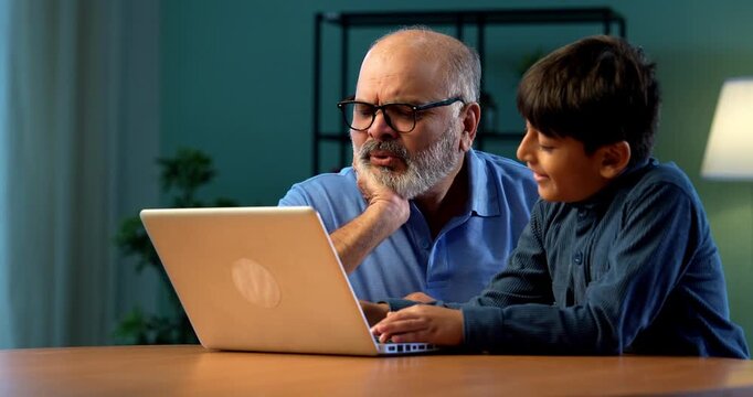Indian grandfather kid using laptop on table in modern home, tech savvy child teaching elderly grandpa how to operate computer, generational learning, family bonding moment of two people indoors