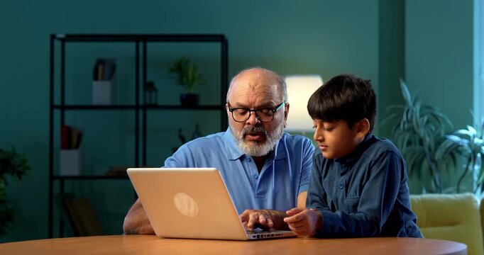 Indian grandfather kid using laptop on table in modern home, tech savvy child teaching elderly grandpa how to operate computer, generational learning, family bonding moment of two people indoors