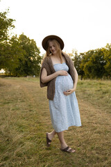 Expecting mother in summer dress and hat standing on a countryside path during golden hour, gently...
