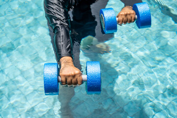 A smiling person in a swim cap and rash guard is submerged in a pool, holding blue aquatic...