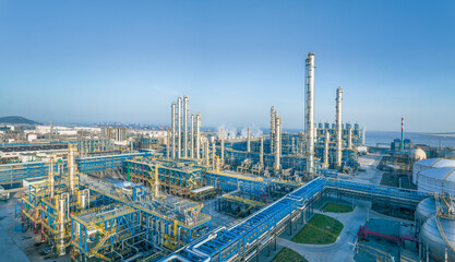 Large modern oil refinery and petrochemical industrial plant with intricate pipelines by the sea under a clear blue sky.