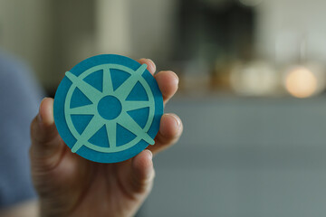 Close-up of a hand holding a teal paper cut-out of a Dharmachakra, a Buddhist symbol of the wheel...