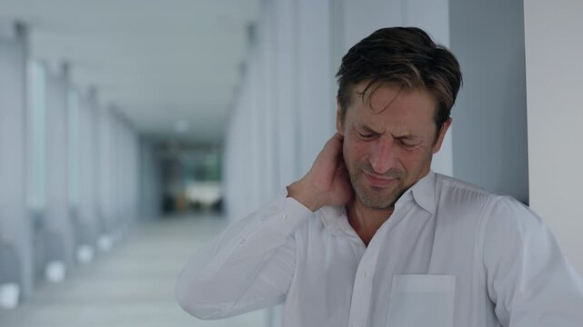 Portrait of businessman in shirt massages neck as feels pain in office hallway. Overworked entrepreneur suffers from muscle crumps caused by work stress in corporate environment