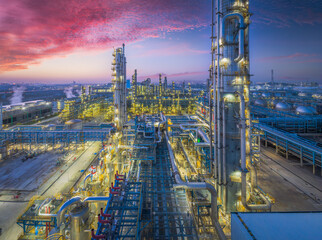 Aerial view of a large illuminated oil refinery and petrochemical industrial plant under a dramatic and colorful sunset sky.