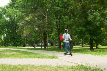 Fototapeta premium Black young man riding electric scooter through park carrying red insulated delivery backpack, moving along paved path surrounded by green trees and grass