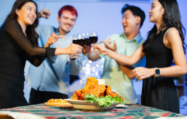 Happy young friends raise red wine glasses in a cozy indoor party setting, laughing and enjoying a festive dinner with good food, warmth, and positive vibes.