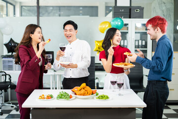 Cheerful coworkers share laughs as one playfully feeds a skewer during a festive office lunch with wine, food, and laughter, celebrating together in a modern workplace.