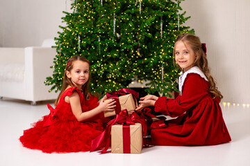 Two little girls, sisters or best friends in red dress sit on a floor under decorated Christmas...