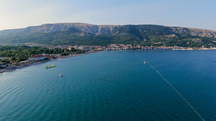 Naklejka premium Baska Beach and Coastline With Clear Turquoise Water, Marina Boats, City Street, Sunny Summer Sunset Aerial View on Krk Island, Croatia Adriatic Sea, Vela Plaza Beach, Mountains and Campsite Cityscape