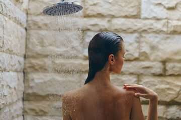 Woman taking outdoor shower with wet hair and stone wall background, enjoying relaxing moment under stream of water in natural setting.