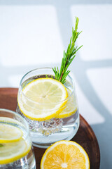 Lemon water with rosemary sprigs and ice cubes served in glasses on round wooden table beside fresh citrus halves