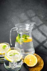 Glass jug of citrus lemonade with mint leaves and ice cubes on dark t summer table in natural sunlight
