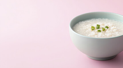 Creamy rice porridge in light green bowl garnished with chopped green onions, set against soft pink background