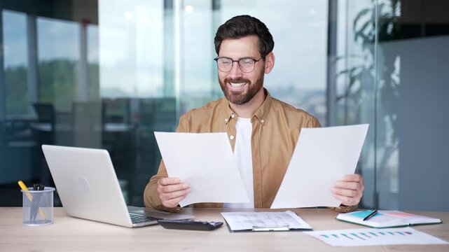A smiling businessman is satisfied with results of a financial report holding documents in his hands sitting at workplace in a business office. Entrepreneur or owner is happy with positive indicators