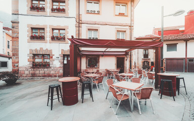 Cozy empty outdoor cafe terrace in a charming European town square, featuring red tables, wicker chairs, and classic architecture with flower-filled window boxes in soft afternoon light