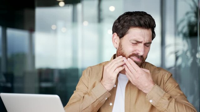 Upset businessman suffering from toothache while sitting at workplace at desk in business office. Male worker holds his hand on his cheek, massaging the sore spot. He needs to see a dentist. Close up