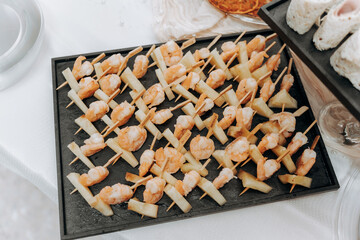 Skewers of shrimp and pineapple are elegantly arranged on a dark tray at a lively event. The...
