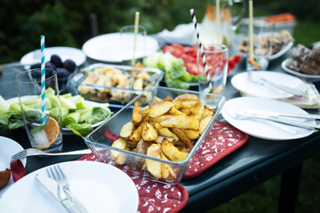 Closeup of crispy baked potato wedges in a glass dish served on outdoor picnic table with plates and fresh vegetables. Concept of homemade food, traditional cuisine, and summer dining. Perfect for