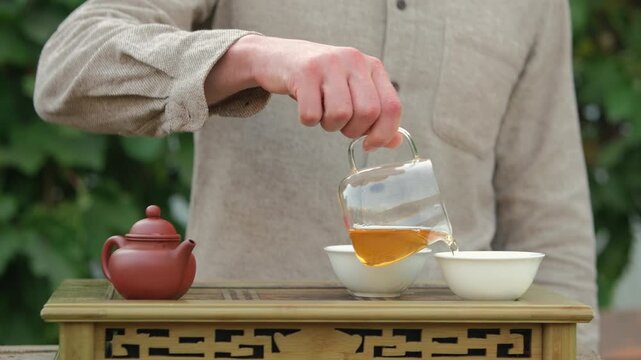 Close up shot of a man standing behind chaban, pouring tea from glass chahai into two white tea cups, part of an outdoor Chinese tea ceremony