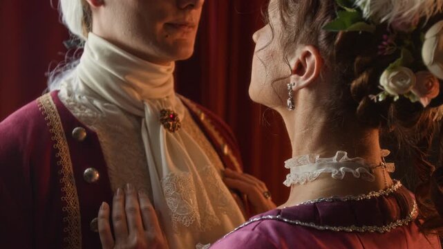 Rear close-up of unrecognizable loving young Victorian couple embracing, woman with flowers in curled hair gently touching chest of young man, dressed in frock coat and lace cravat with brooch