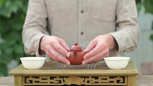 Close up shot of a man standing behind chaban, putting Chinese clay tea pot closer to the camera pointing toward it with two hands, two white tea cup on eather side of a tea board.
