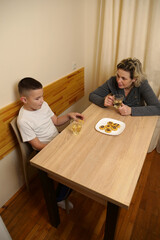 Family moment with mother and son sitting at table, drinking tea, eating cookies, and enjoying warm conversation together indoors.