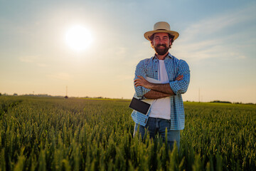 Happy farmer crossing arms while holding a tablet in a lush green wheat field during sunset, relishing the success of agricultural business and the promise of a bountiful harvest