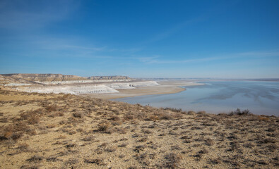 Expansive desert landscape with rugged terrain, white limestone cliffs, and a serene reflective water body under a clear blue sky, located in a remote arid region.