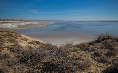 A vast salt plain with white cliffs and reflective water under a clear blue sky, surrounded by barren desert terrain. Located in Mangystau, Kazakhstan.