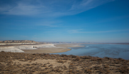 The expansive and arid landscape of the Ustyurt Plateau in Kazakhstan, with a large salt flat. The terrain is barren, with low, eroded cliffs visible in the distance under a vast blue sky.
