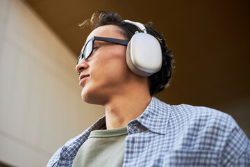 Young adult Asian man wearing over ear headphones and glasses looking away from camera outdoors, listening to music or podcast, casual expression, short wavy hair visible