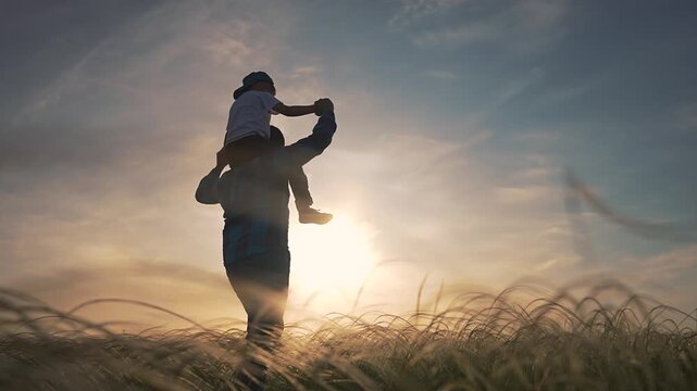 Child sitting on father shoulders in field. Father holding son hands. Boy smiling while dad walking in grass. Parent playing with kid under sunset. Father and child enjoying evening walking time