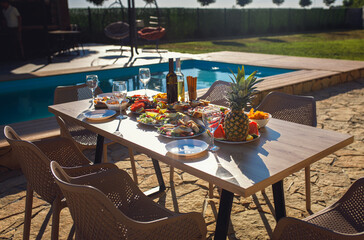 Outdoor dining table with food, wine and fruits set by swimming pool in backyard.