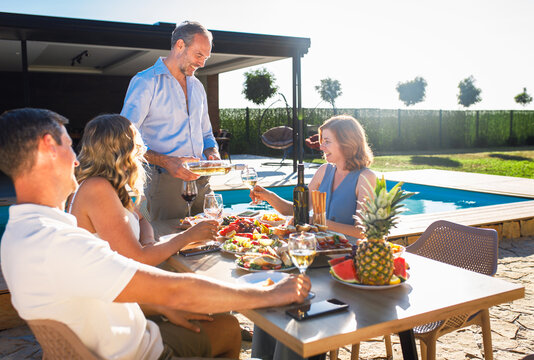 Group of adult friends having lunch by swimming pool in sunny backyard.