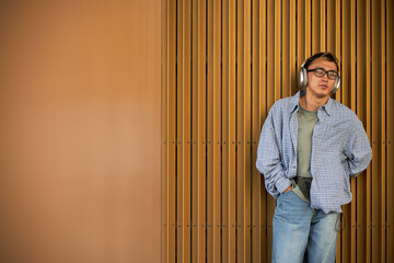 Young adult Asian man standing against vertical wooden slats, wearing headphones and glasses,...