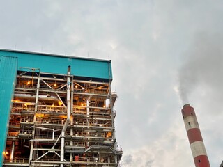 Industrial complex with soaring red and white smokestack releasing steam against a clouded sky...