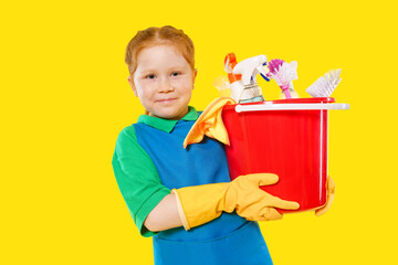 Young child dressed in cleaning gear holds a bucket filled with cleaning supplies against a bright yellow backdrop