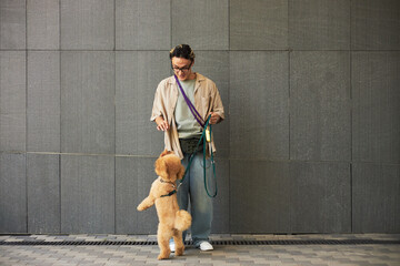 Young adult Asian man standing outdoors interacting with fluffy dog on leash, holding treat while dog standing on hind legs, casual urban setting, textured wall background