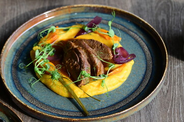 Roasted lamb served with creamy pumpkin purée and seasonal vegetables, a colorful autumn dish presented on a rustic plate and wooden table in a restaurant setting.