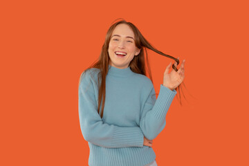 Joyful young woman playfully twirls her hair while smiling against a vibrant orange background during a casual indoor moment
