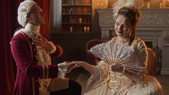 Medium shot of noble young Caucasian gentleman in red surcoat, breeches, wig standing on knee in front of aristocratic lady, kissing her hand, confessing love while proposing marriage