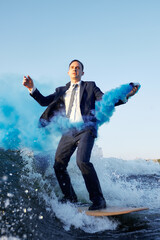 Young man surfer in classic suit with blue smoke bomb in his hand rides on the wave. Happy clerk...